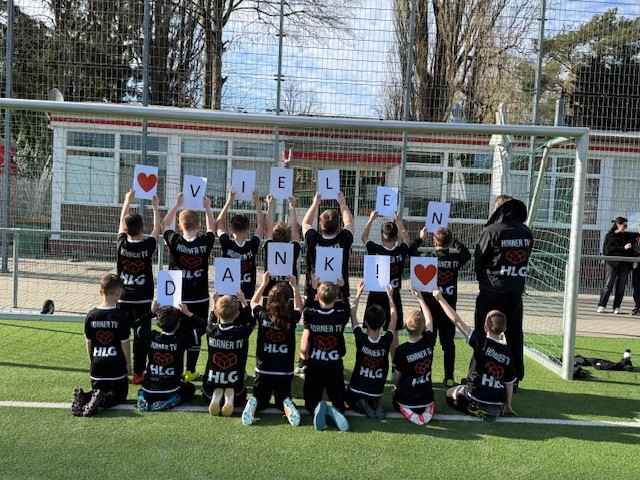 Kinder in Sporttrikots halten große Buchstaben, die „VIELEN DANK!“ bilden, vor einem Fußballtor.